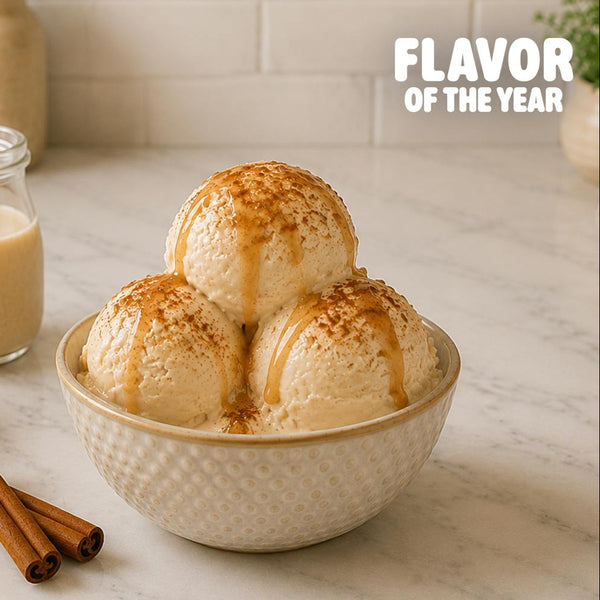 Three scoops of vanilla ice cream in a textured cream-colored bowl, topped with caramel drizzle and cinnamon dusting, set on a marble countertop with a glass jar of vanilla extract, a small bowl of cinnamon powder, and cinnamon sticks. The text 'FLAVOR OF THE YEAR' is visible in the top right corner.