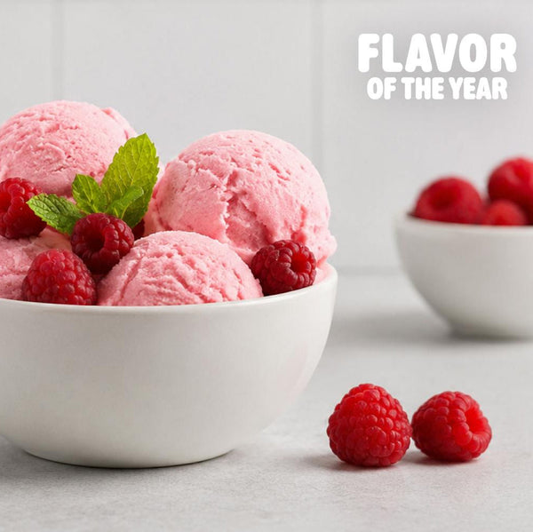 Scoops of pink raspberry ice cream in a white bowl, garnished with fresh raspberries and a mint leaf, with additional bowls of raspberries blurred in the background and the text 'FLAVOR OF THE YEAR' in the top right.